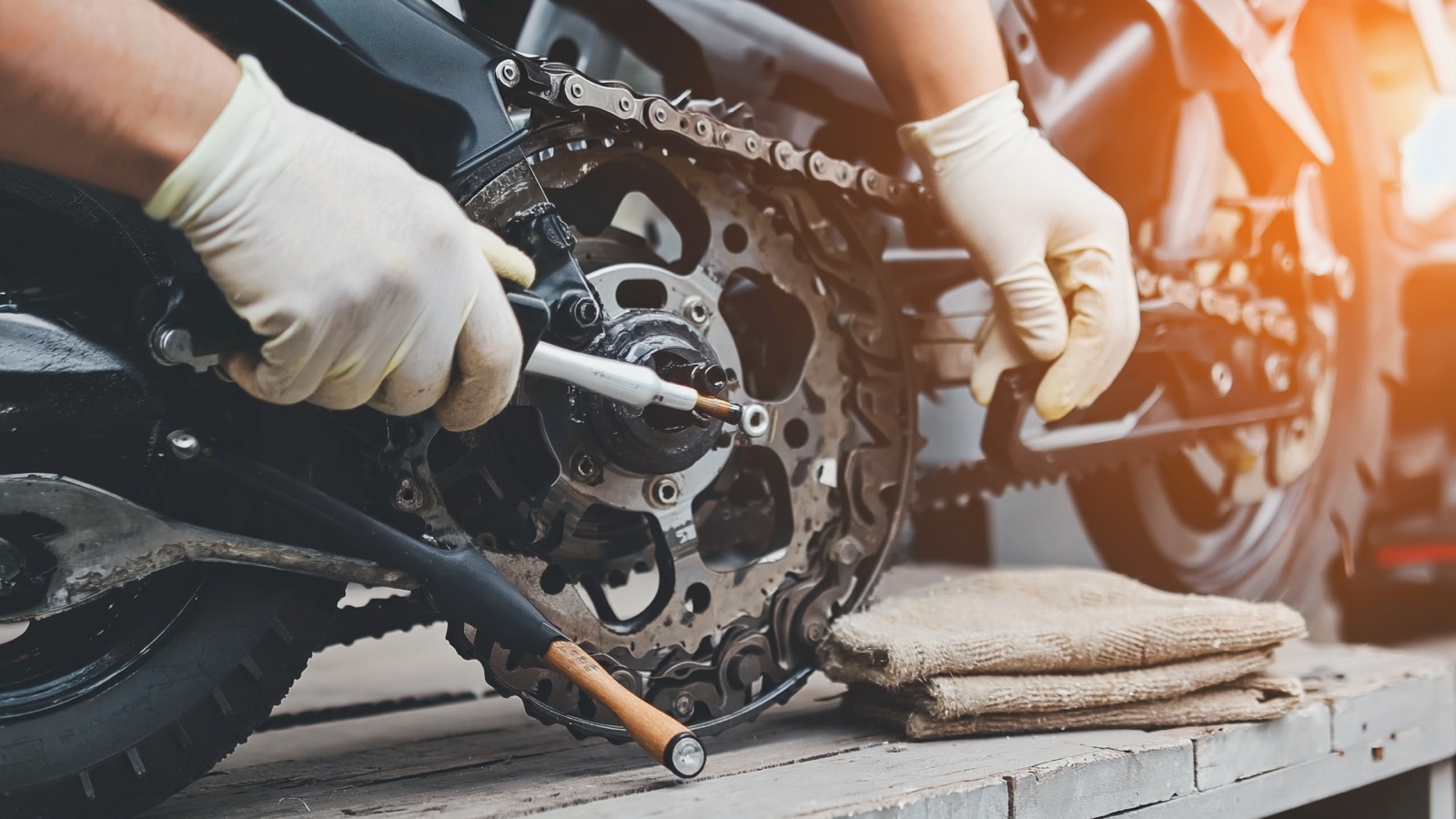 Behandschuhte Hände ölen im warmen Morgenlicht die Antriebskette und das hintere Kettenrad eines Motorrads, daneben ein Lappen und eine kleine Bürste.