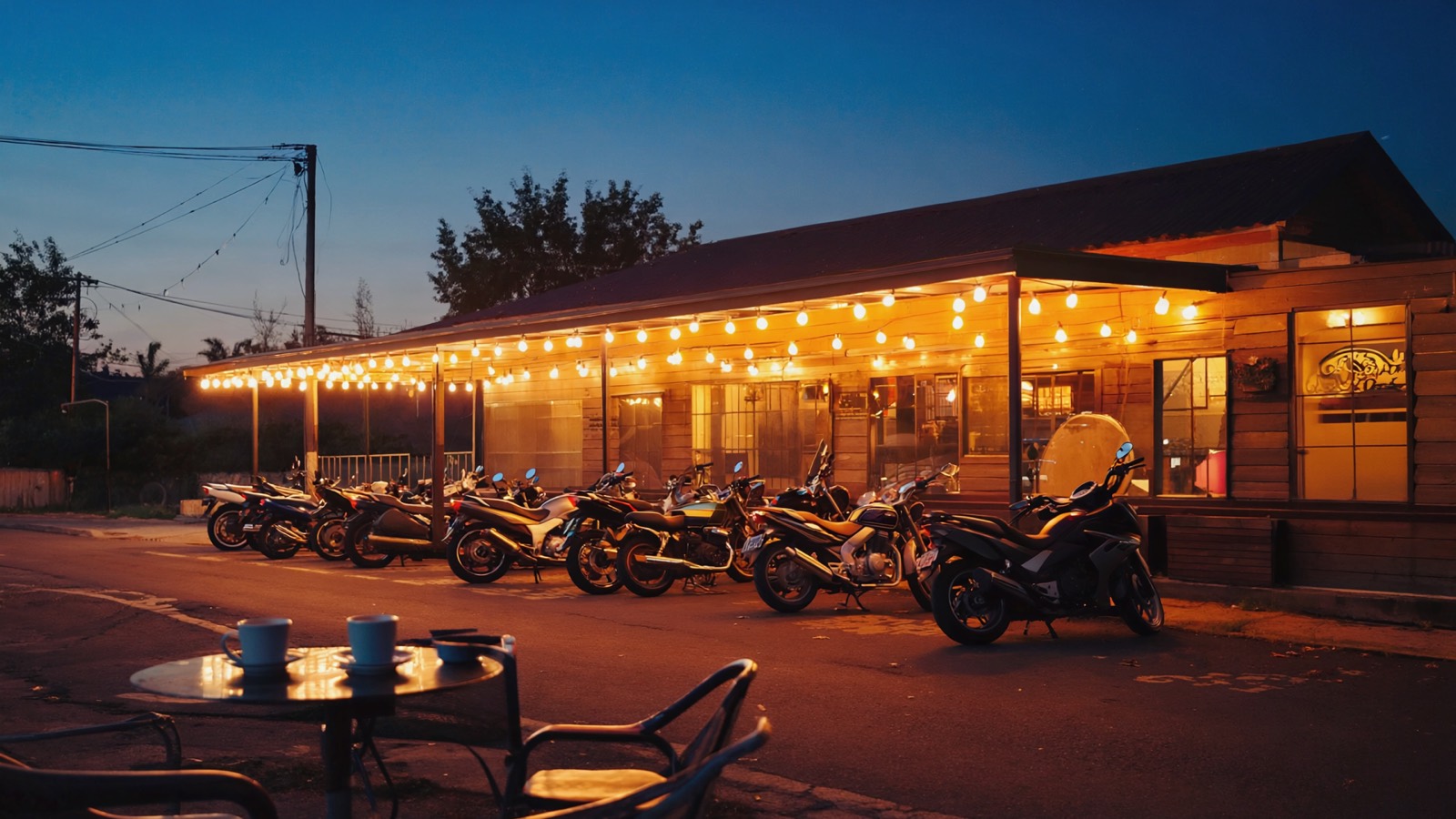Mehrere geparkte Motorräder vor einem Straßencafé in der Abenddämmerung, warme Lichterketten glühen, Kaffeetassen auf einem Tisch, entspannte Treffpunkt-Atmosphäre.